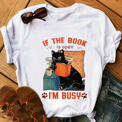 white t-shirt with a black catleaning on a table top with a book in its hand and a cup of coffee on the table top, words on t-shirt say, If the book is open, (picture of paw print) I'm busy (picture of paw print)
